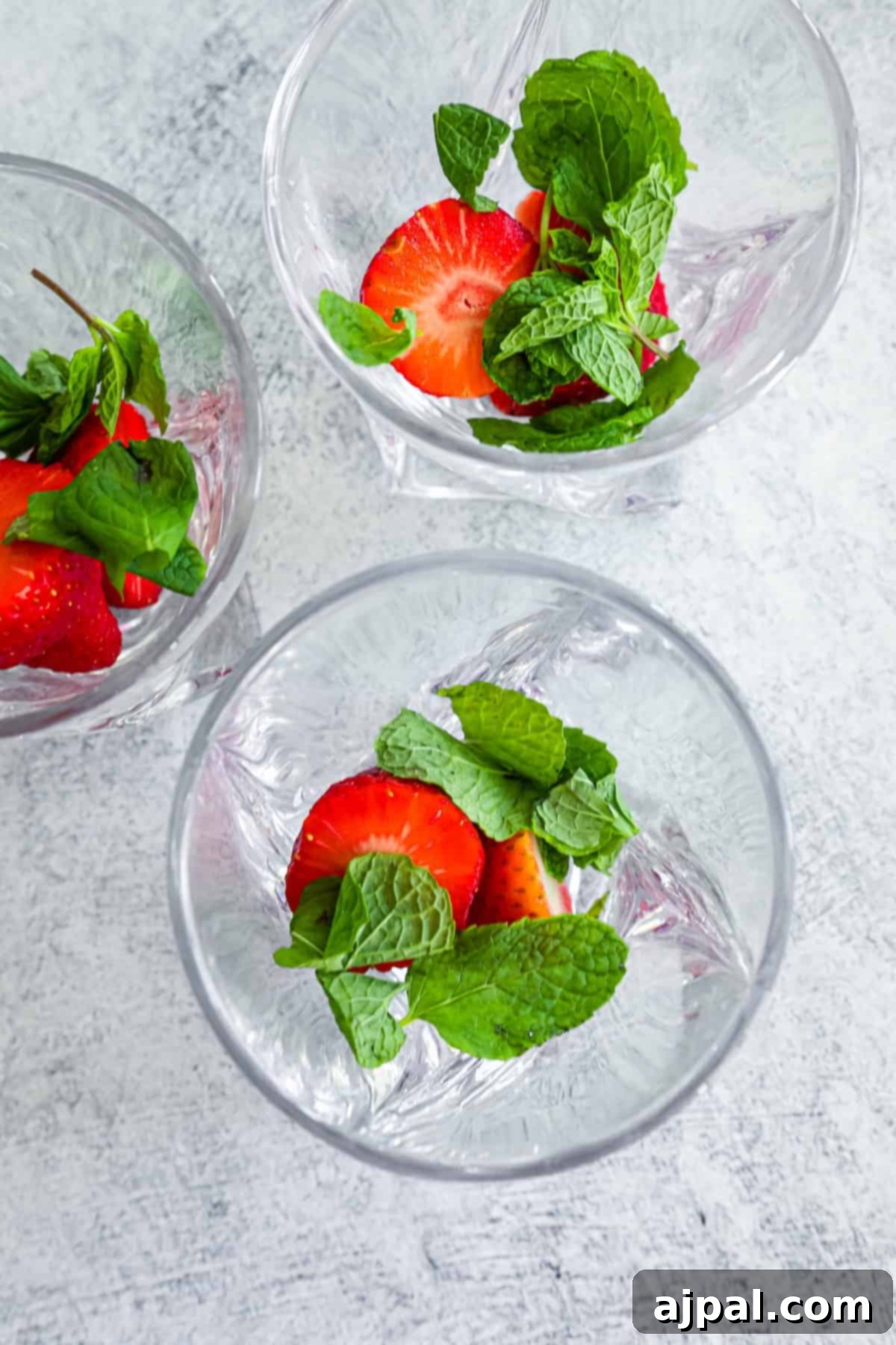 Three crystal bowls filled with water, fresh strawberries, and mint leaves on a textured surface, emphasizing fresh ingredients.