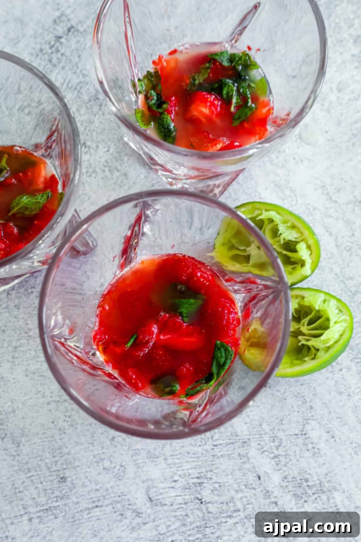 Top view of three glasses containing remnants of muddled red fruit with bits of basil and mint, accompanied by lime halves on a textured gray surface, showing the muddling step.