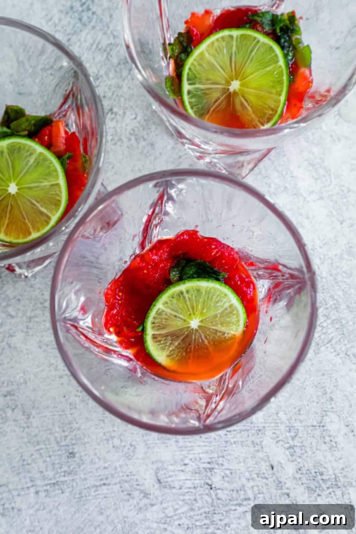 Three cocktail glasses on a textured surface, each containing a bright red muddled fruit mixture topped with a lime slice and mint leaves, preparing for the liquid.