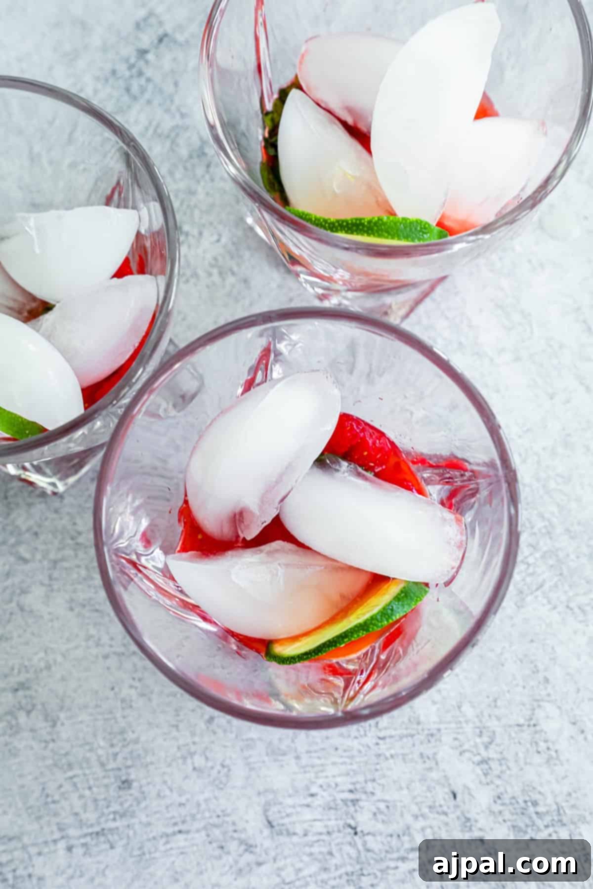 Three glasses containing ice cubes, muddled fruit, and lime slices on a textured gray surface, ready for sparkling water.