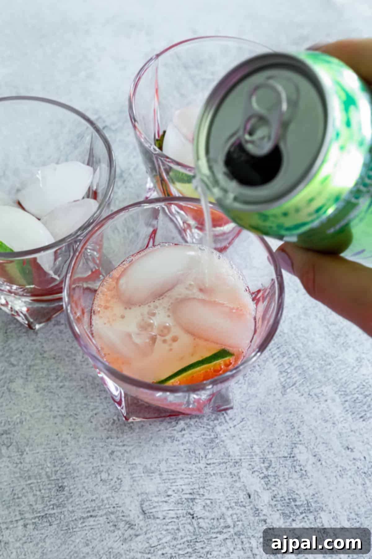 Pouring a clear carbonated beverage into a glass containing ice cubes, muddled strawberries, and a slice of lime, set on a textured gray surface, capturing the fizz.