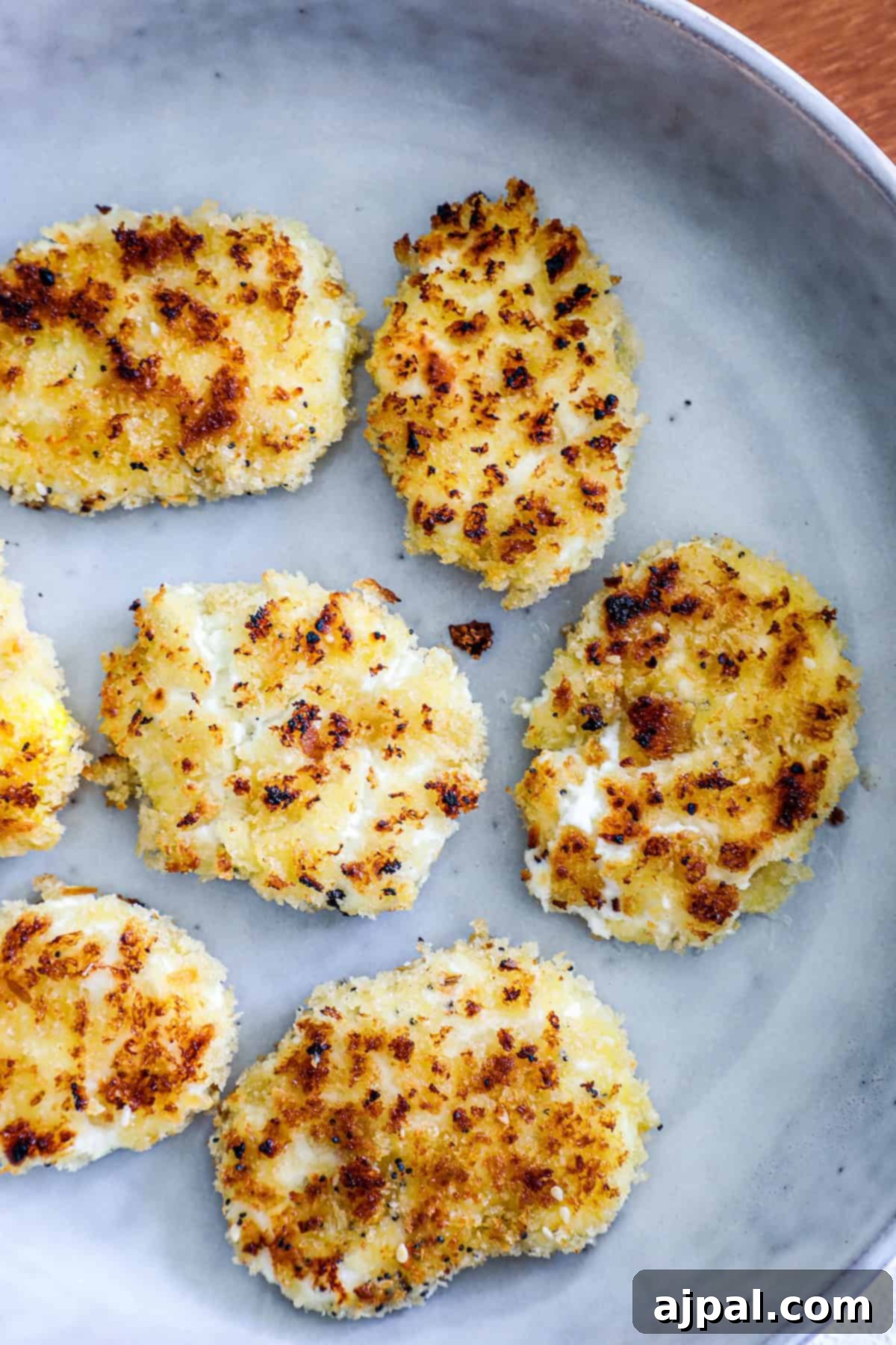 Six golden-brown fried goat cheese patties are artfully arranged on a large ceramic plate, highlighting their crispy texture.