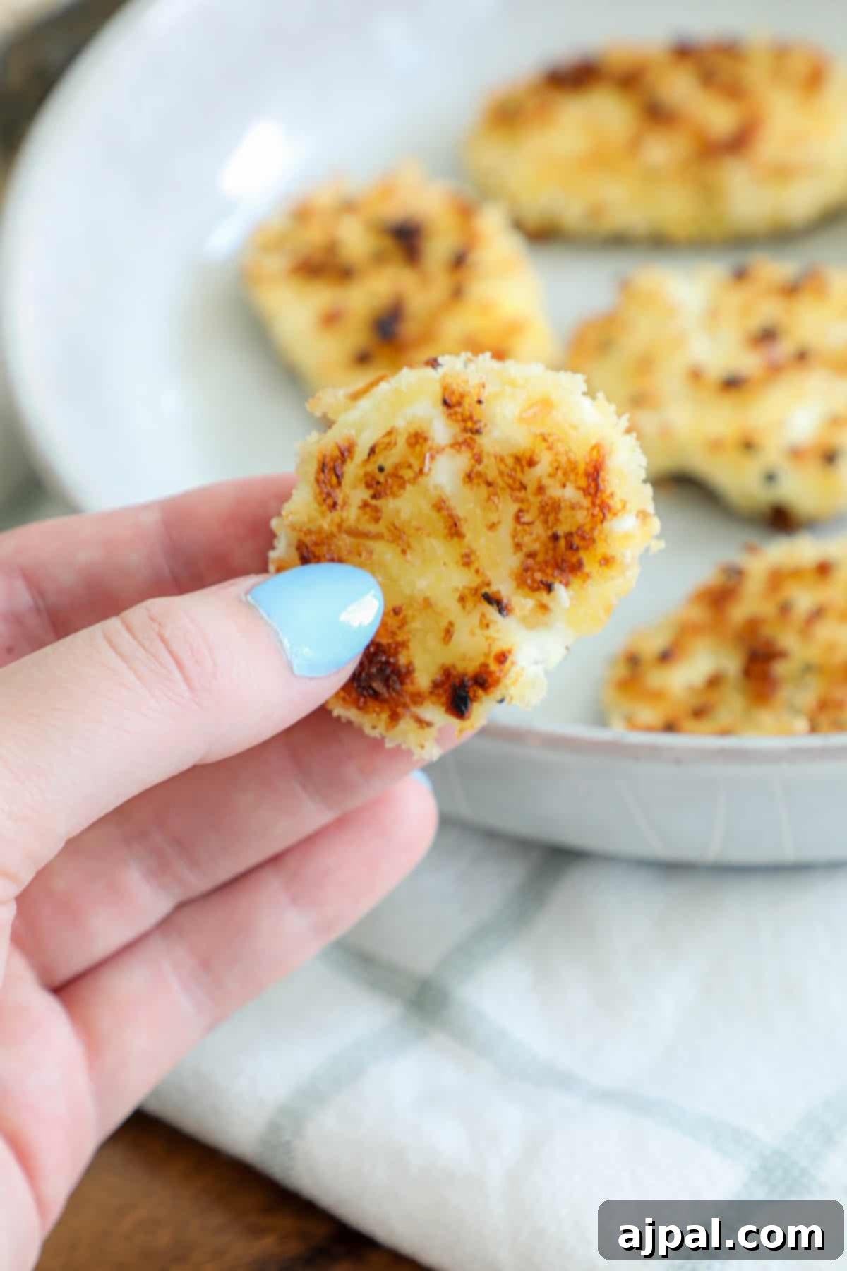 A hand with blue nail polish holds a small, round, golden-brown fried goat cheese patty, hovering above a plate of similar perfectly cooked patties.
