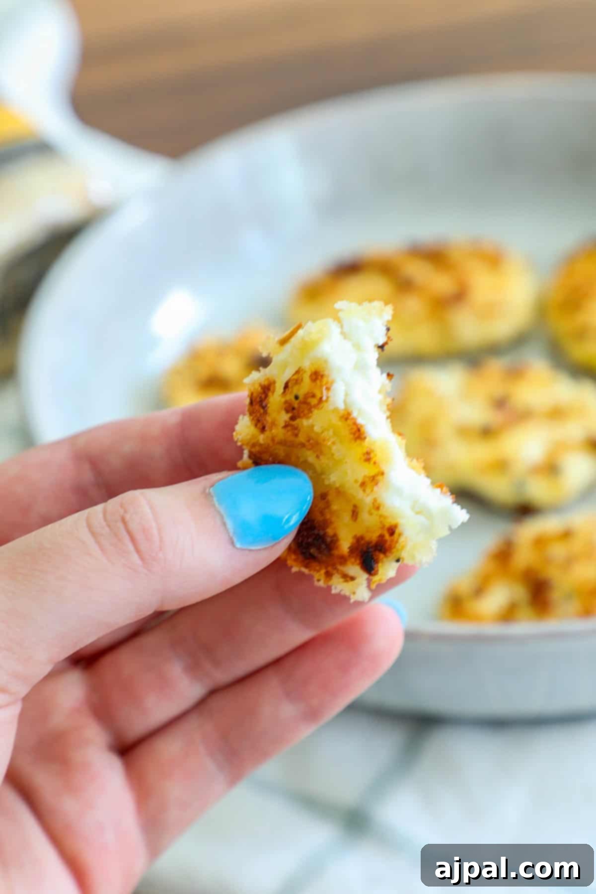 A hand gently holds a single golden-brown fried goat cheese bite, showcasing its crunchy coating, with a plate of more bites visible in the background.