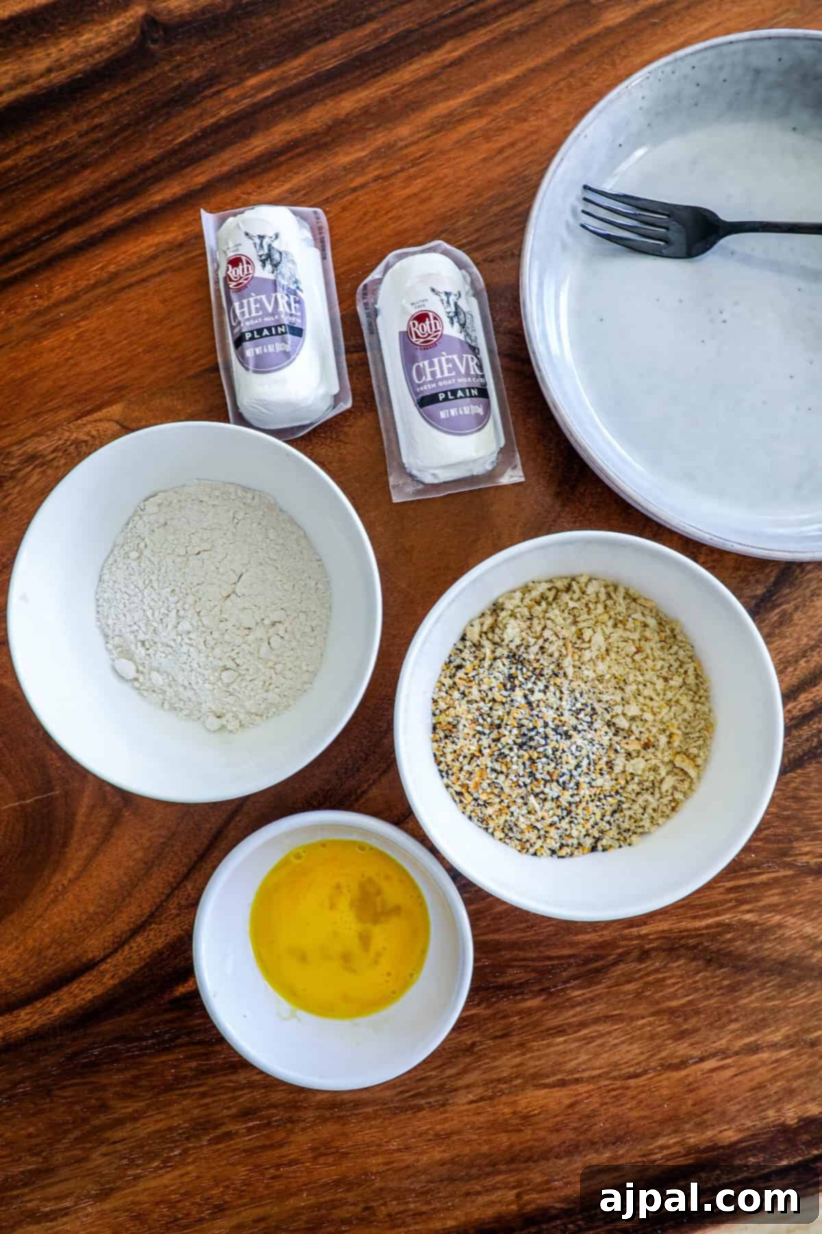 Recipe ingredients, including bowls of flour, panko breadcrumbs, a whisked egg, and seasoning containers, are artfully arranged on a wooden table.