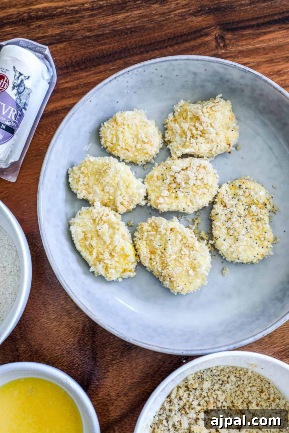 Uncooked breaded chicken pieces rest on a gray plate, with separate bowls of breadcrumbs and flour alongside a bag of batter mix on a wooden table, preparing for frying.
