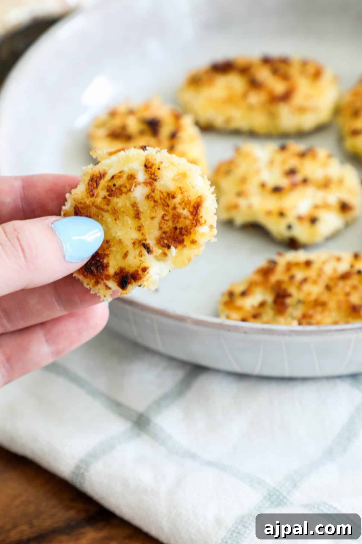 A person holding a golden-brown, breaded cauliflower bite with more pieces on a plate in the background, ready to be served.