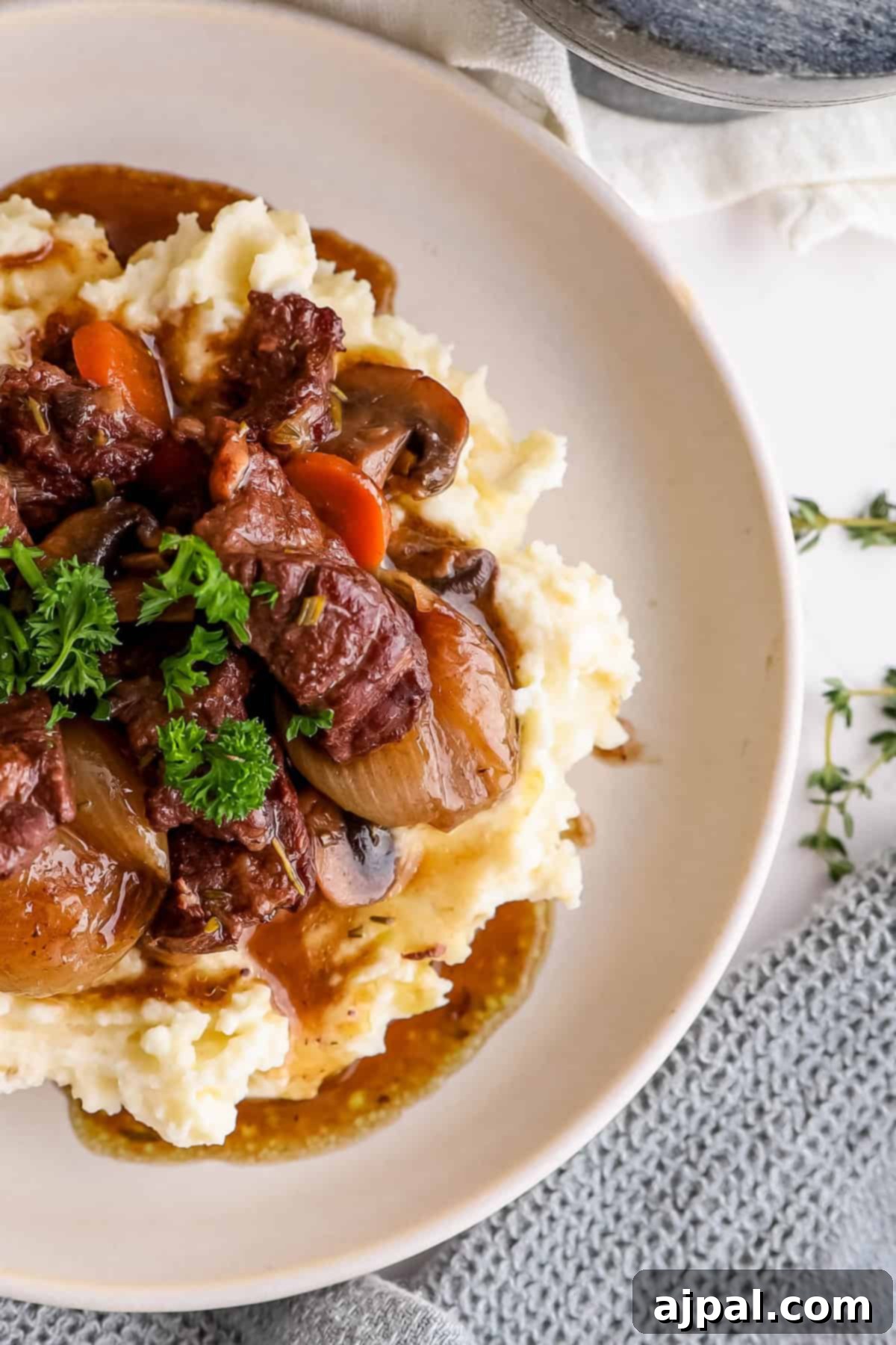 Hearty Burgundy Beef Stew 2 A plate of beef stew served over mashed potatoes, garnished with parsley, accompanied by thyme on a light background.