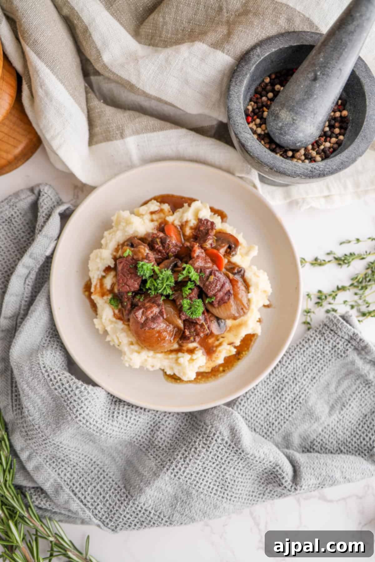 A delectable plate of Beef Bourguignon served over creamy mashed potatoes, garnished with vibrant fresh herbs. Beside the plate, a mortar filled with fragrant peppercorns and sprigs of thyme adds a rustic, aromatic touch to the culinary display.
