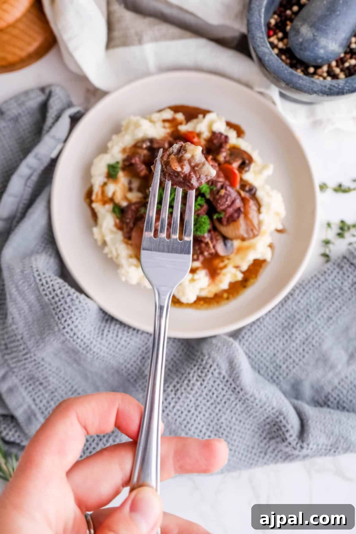A person elegantly holds a fork with a succulent piece of beef, poised over a inviting plate of Beef Bourguignon and creamy mashed potatoes. The rustic kitchen setting creates a warm and authentic backdrop for this delectable meal.