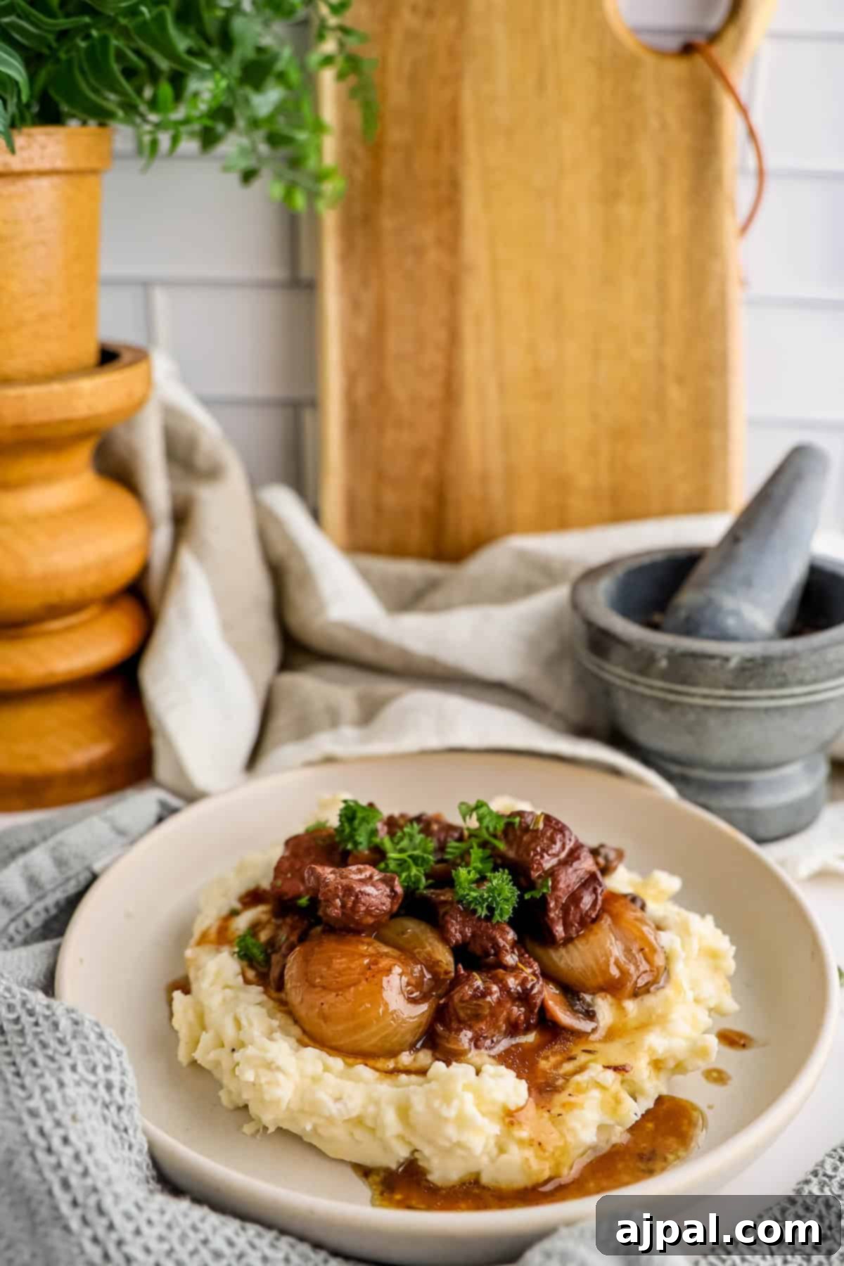 A delightful plate of Beef Bourguignon featuring tender beef and pearl onions, served over creamy mashed potatoes, garnished with fresh parsley. A wooden pepper mill and mortar sit elegantly on the kitchen counter beside it, adding to the rustic culinary scene.