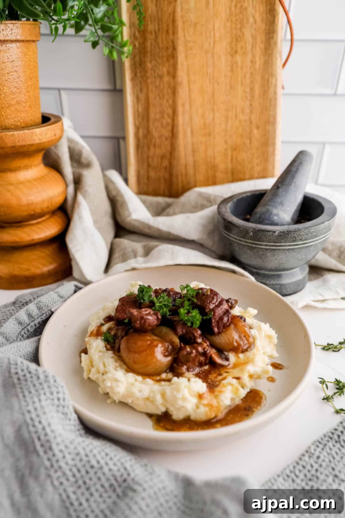 Hearty Burgundy Beef Stew 3 A plate of beef stew with whole mushrooms served over mashed potatoes, garnished with parsley, set on a wooden kitchen counter.