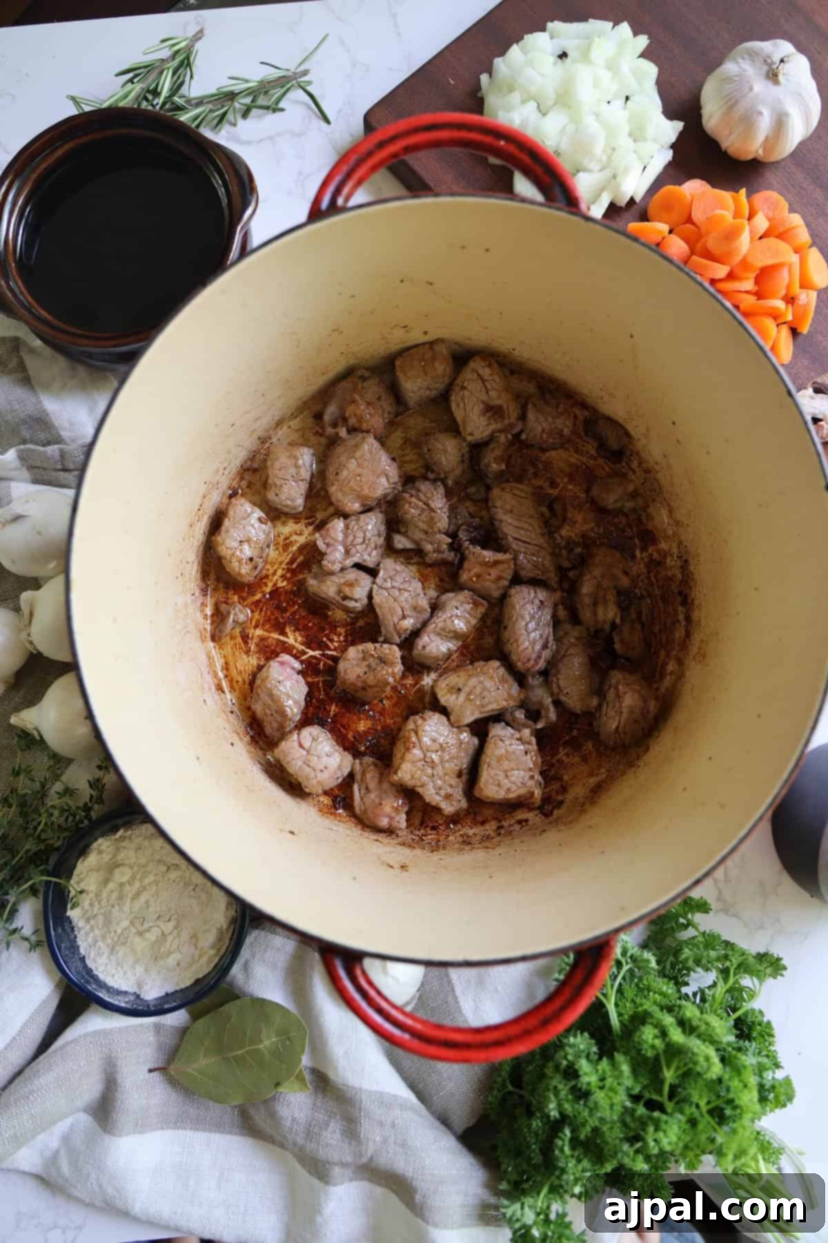A vibrant top-down view of a red cooking pot on a stove, containing perfectly browned beef pieces. Surrounding the pot are fresh ingredients like sliced carrots, diced onions, minced garlic, and an assortment of fragrant herbs, all prepped for the next steps.
