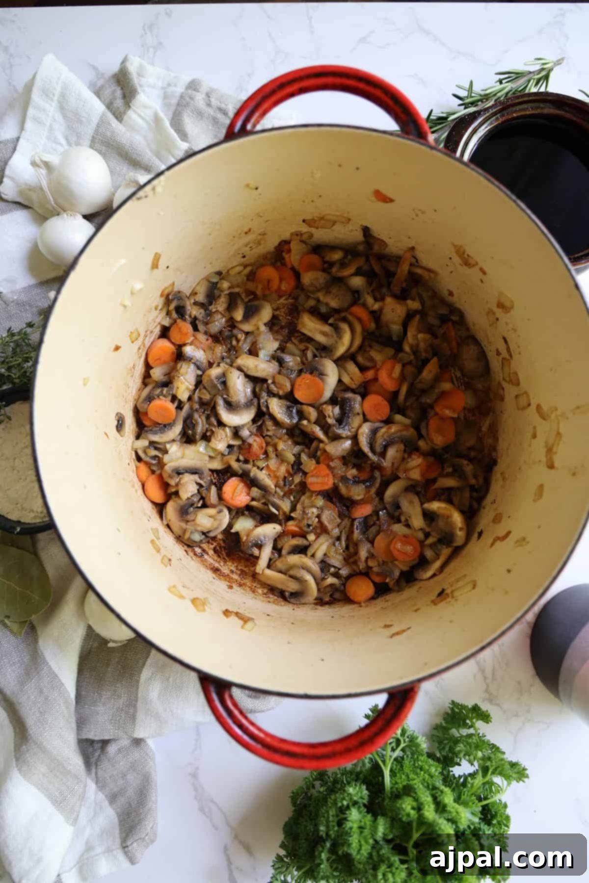 Hearty Burgundy Beef Stew 7 A large red pot containing sautéed mushrooms and sliced carrots on a kitchen counter, surrounded by cooking ingredients and utensils.