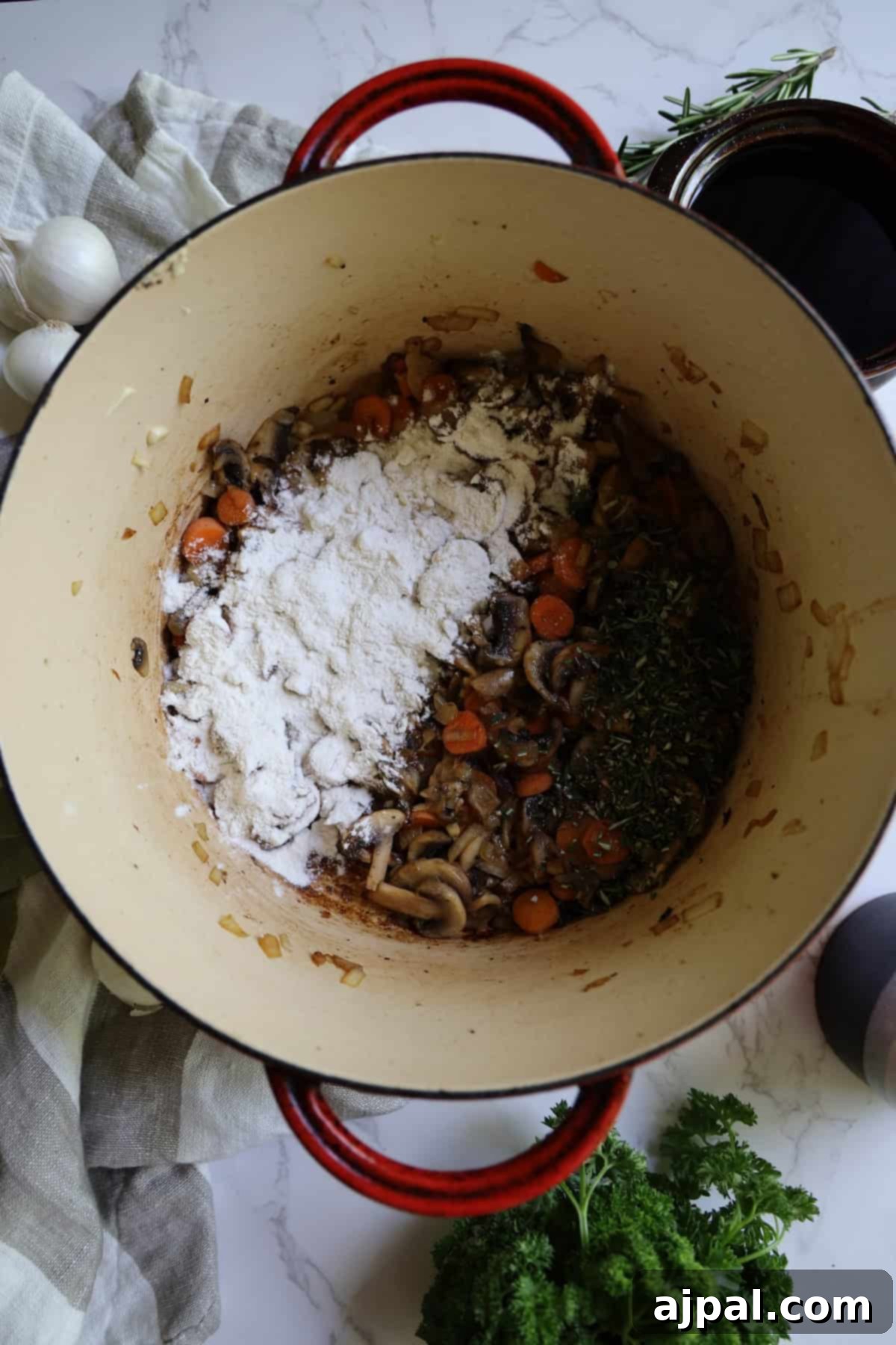 A top-down perspective reveals a cooking pot on a kitchen counter, holding a mix of flour, fragrant herbs, tender mushrooms, and bright sliced carrots. Various scattered ingredients and spices surround the pot, setting the stage for a culinary creation.