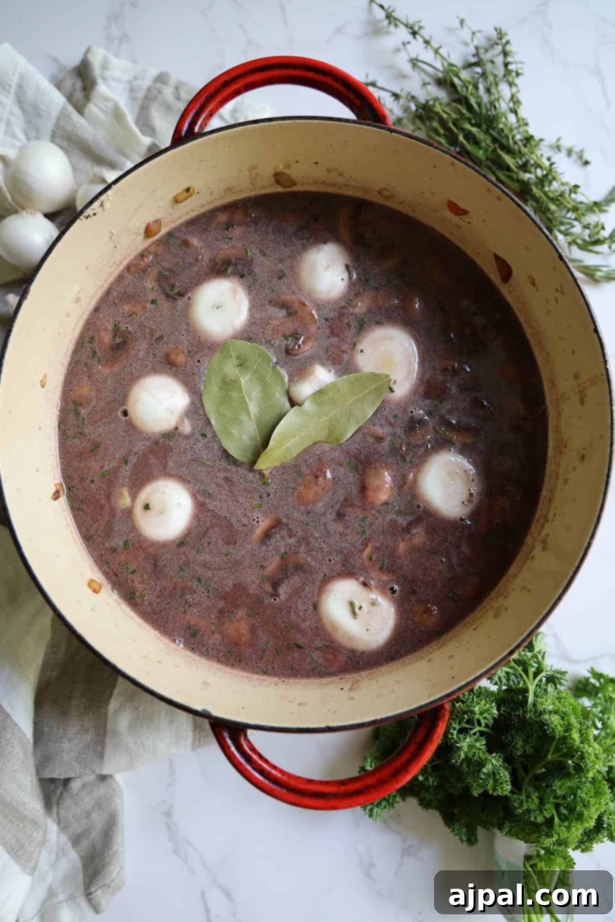 Hearty Burgundy Beef Stew 9 A red enameled pot filled with black bean soup, garnished with whole onions, bay leaves, surrounded by garlic, thyme, and parsley on a marble countertop.