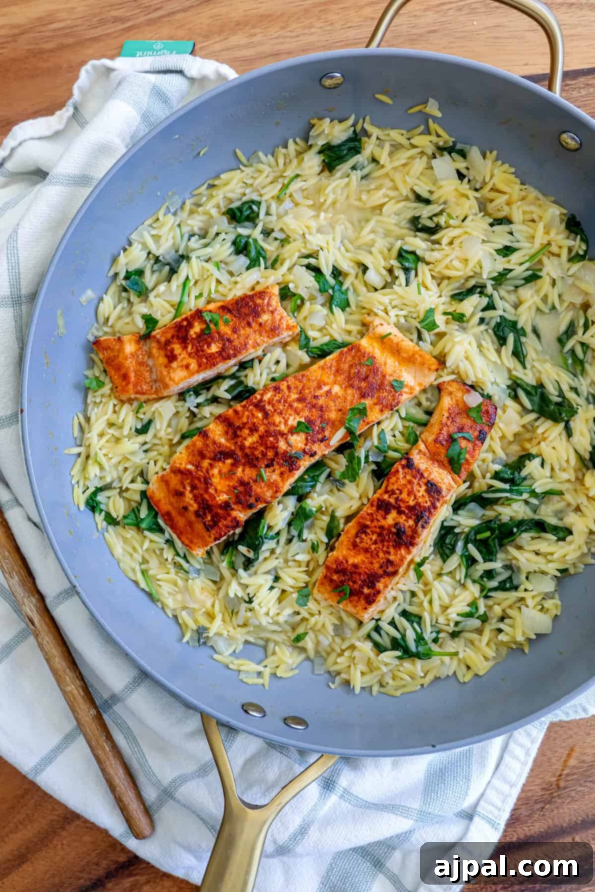 Creamy Salmon Florentine Orzo 3 A skillet containing orzo pasta with spinach and three pieces of grilled salmon on top, placed on a wooden surface.