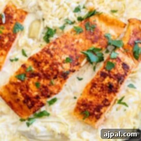 Three pieces of seasoned, grilled salmon are served over a bed of creamy orzo pasta with artichokes, garnished with chopped parsley.