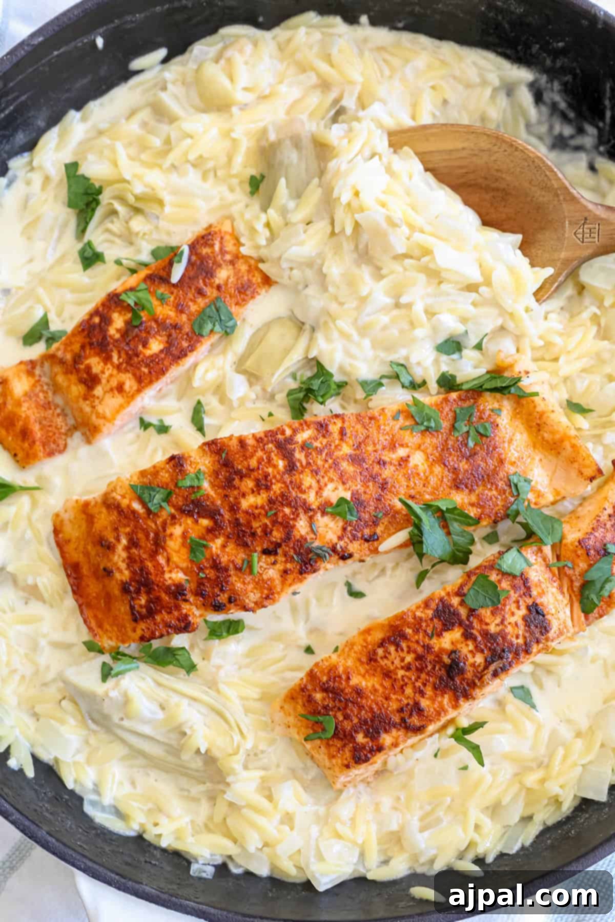 A large skillet holding creamy artichoke orzo pasta with two salmon fillets cooked on top, garnished with herbs.