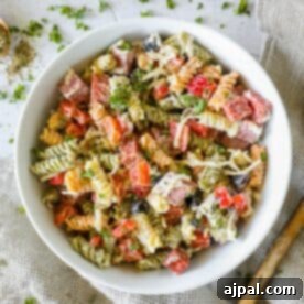 A bowl of the best Italian pasta salad featuring tricolor rotini mixed with diced red bell peppers, black olives, chopped parsley, and shredded cheese, set on a textured cloth background.