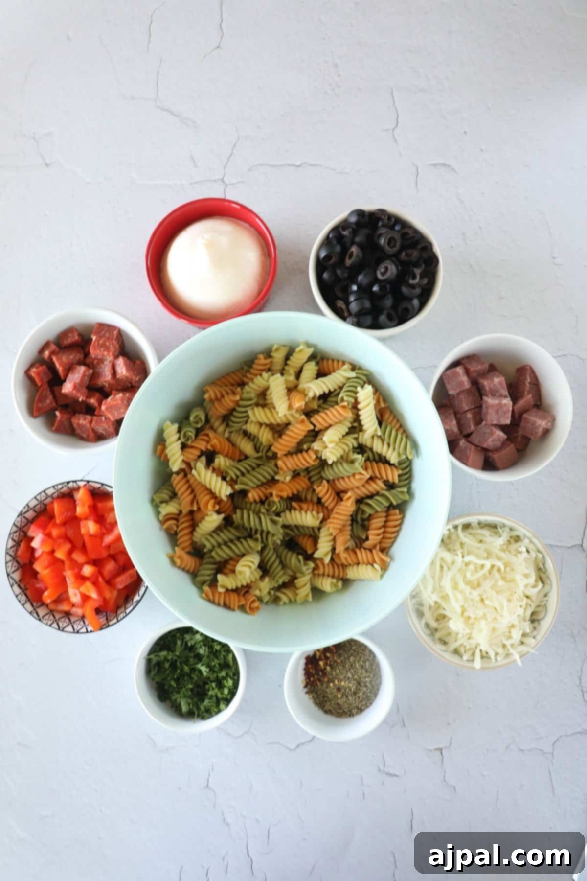 Creamy Italian Pasta Salad 5 Fresh ingredients laid out on a wooden board, including a bowl of cooked rotini pasta, diced red bell pepper, shallots, black olives, salami, and mozzarella, ready for assembly into a creamy Italian pasta salad.