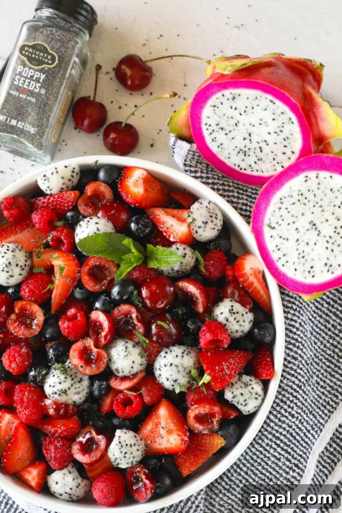 Sweet Berry Burst 2 A bowl of mixed fruit including strawberries, blueberries, raspberries, and dragon fruit, garnished with fresh mint leaves. There are also fresh cherries, dragon fruit halves, and a jar of poppy seeds nearby, highlighting the fresh ingredients for this delicious berry and cherry fruit salad.