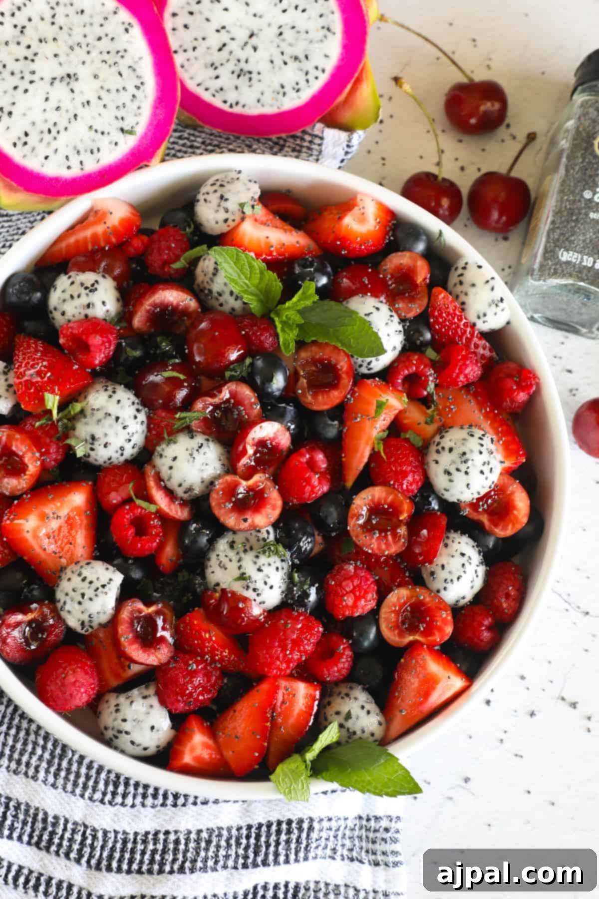 Sweet Berry Burst 7 A bowl of mixed fruit salad containing strawberries, dragon fruit, cherries, blueberries, and raspberries, garnished with mint leaves. Two halves of a dragon fruit and some cherries are beside the bowl, ready for a healthy treat.