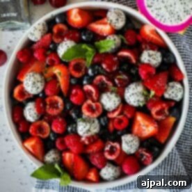 A white bowl filled with a colorful mix of strawberries, blueberries, raspberries, cherries, and sliced dragon fruit, garnished with fresh mint leaves, placed on a blue and white cloth.