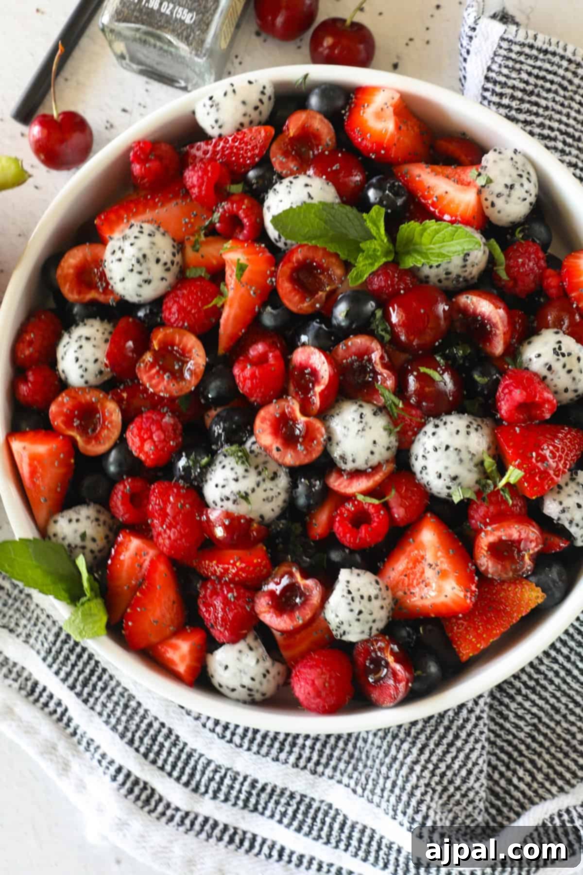 Sweet Berry Burst 3 A white bowl filled with a mixed fruit salad featuring strawberries, raspberries, blueberries, cherries, and dragon fruit pieces, garnished with mint leaves, ready to be served as a refreshing dessert or side dish.