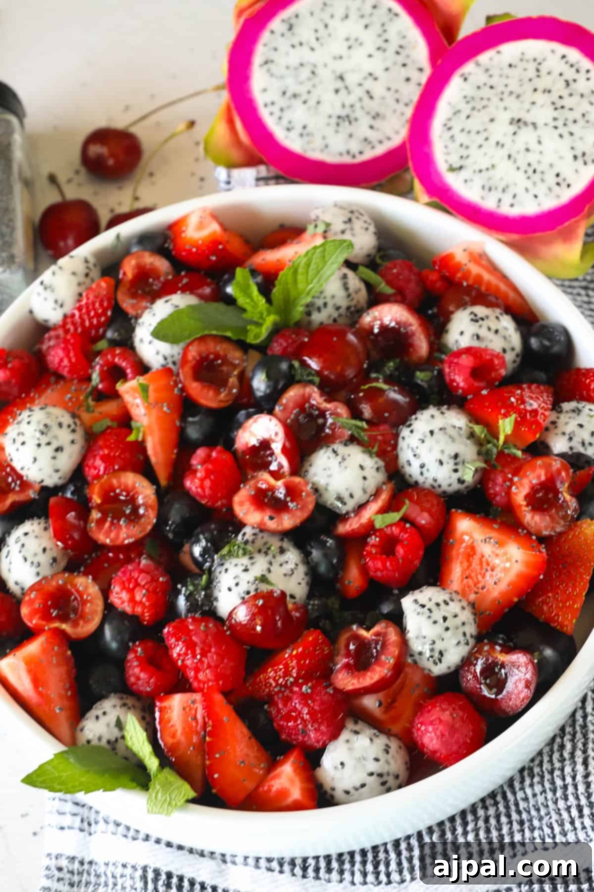 Sweet Berry Burst 5 A bowl filled with a mixed fruit salad, including sliced strawberries, raspberries, blueberries, cherries, and dragon fruit, garnished with mint leaves. A halved dragon fruit is in the background, showcasing the fresh, vibrant ingredients.