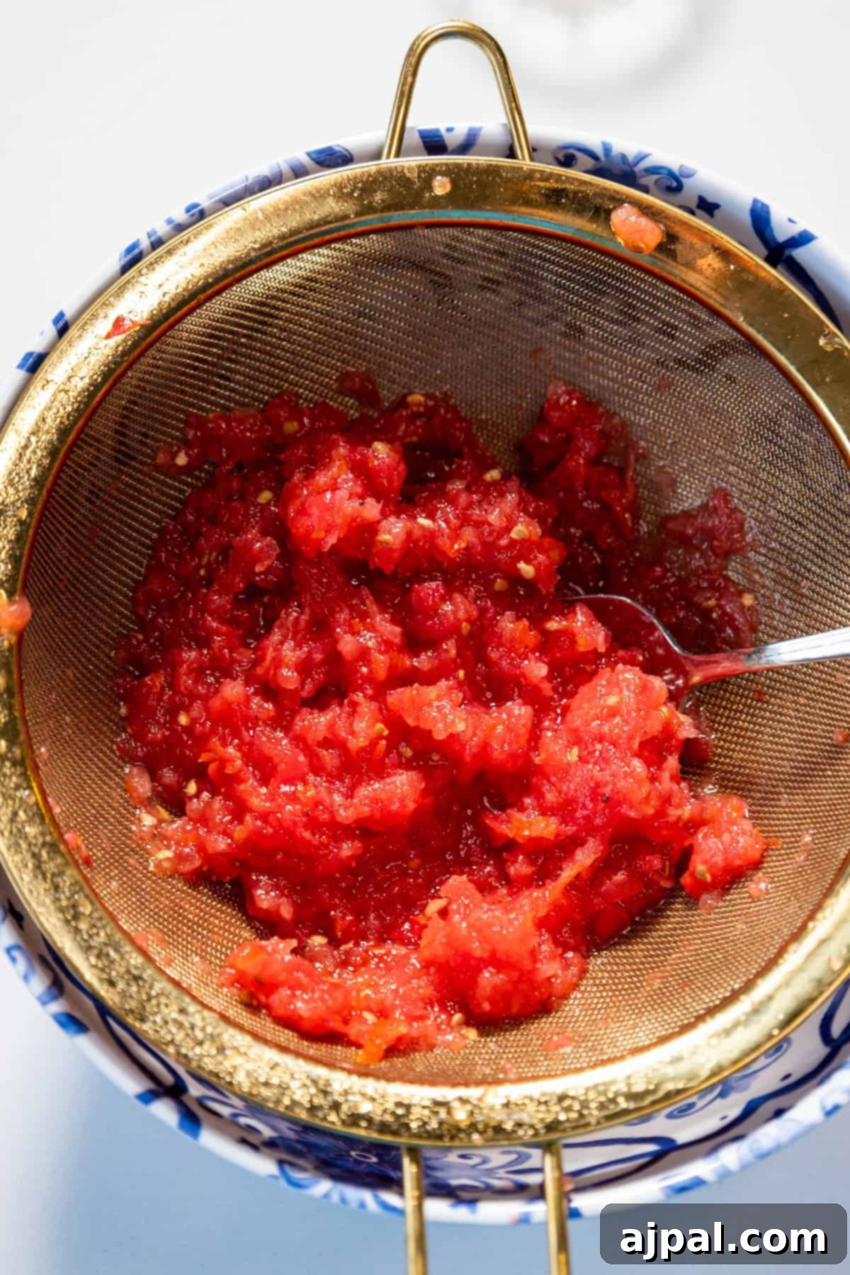 Grated tomato pulp in a strainer, seasoned with salt and spices, being lightly stirred.