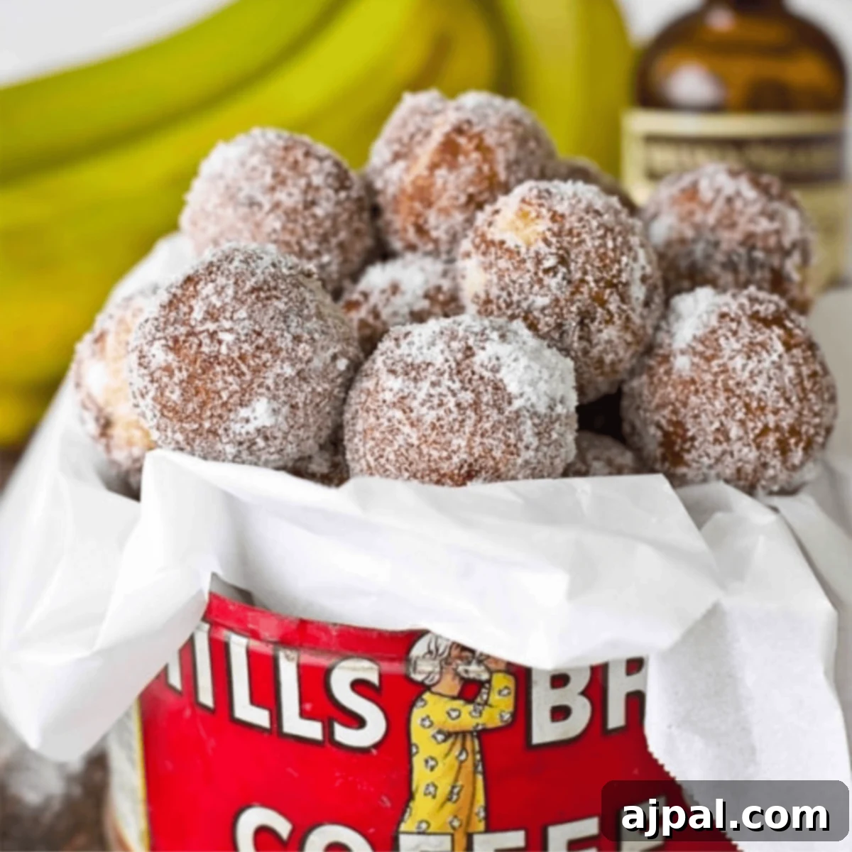 A container filled with powdered sugar-coated banana bread donut holes, surrounded by fresh bananas and a bottle in the background, evoking a tempting quick treat.