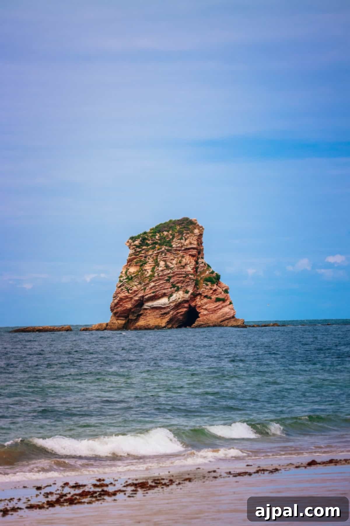 Hondarribia Uncovered 11 A rocky island with vegetation sits in shallow ocean water under a clear blue sky, with gentle waves breaking on a beach in the foreground.