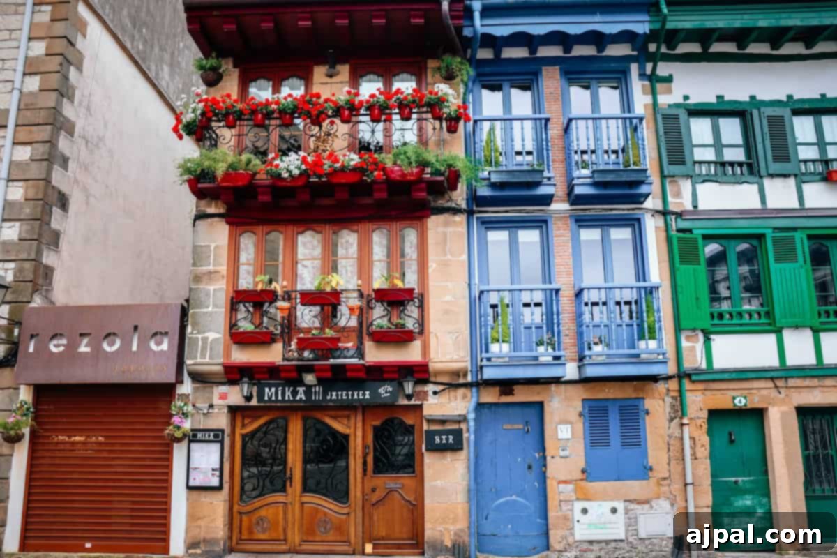 Hondarribia Uncovered 12 Colorful, narrow European buildings with wooden balconies and flowerboxes line a street. A shop named "MIKA" is on the ground floor of the red building.
