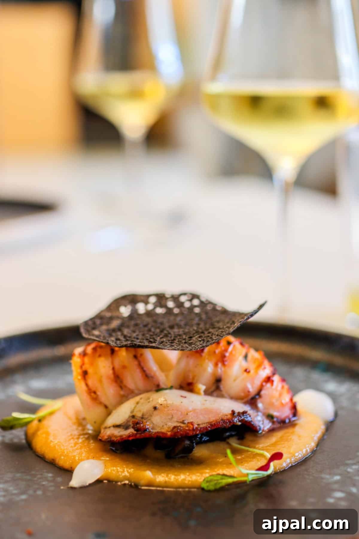Hondarribia Uncovered 13 A gourmet dish featuring grilled seafood atop a creamy sauce, garnished with herbs and a crisp black tuille, with two glasses of white wine in the background.