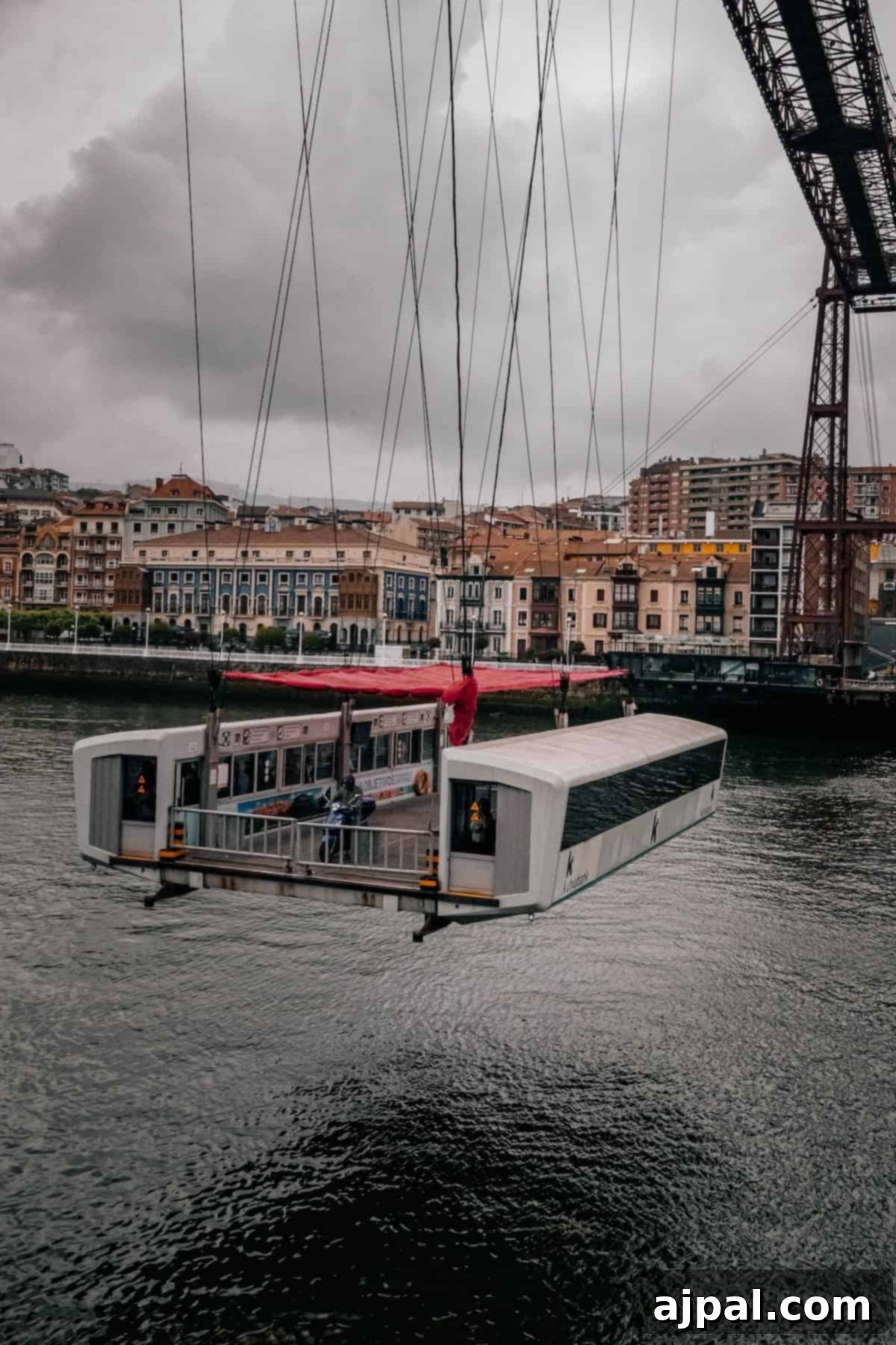 A suspended gondola is transporting a vehicle and passengers across a river, with an urban landscape and overcast sky in the background.