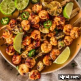 Margarita grilled shrimp skewers garnished with lime wedges, sliced green peppers, and charred lemon halves on a beige plate.