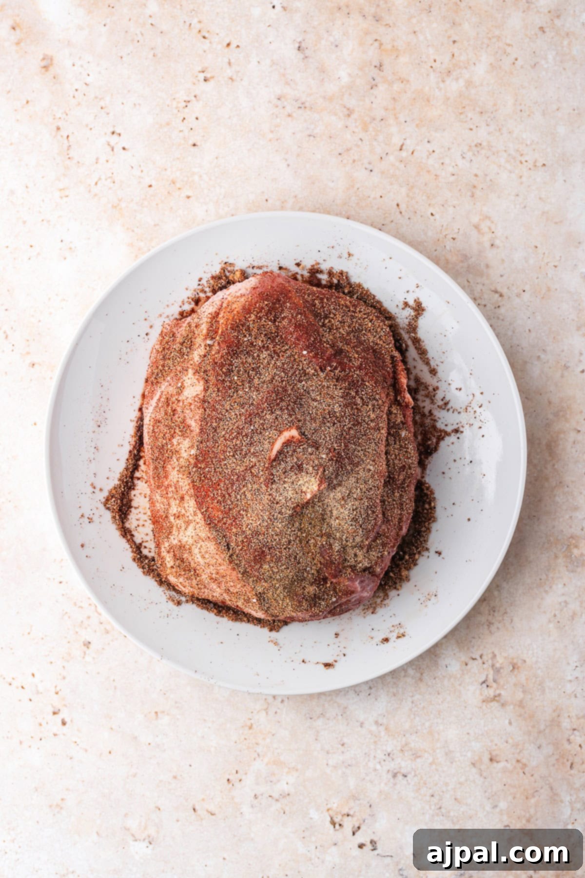 Seasoned raw beef, perfect for your Dutch oven, sits on a white plate against a textured beige surface, ready to transform into a flavorful delight.