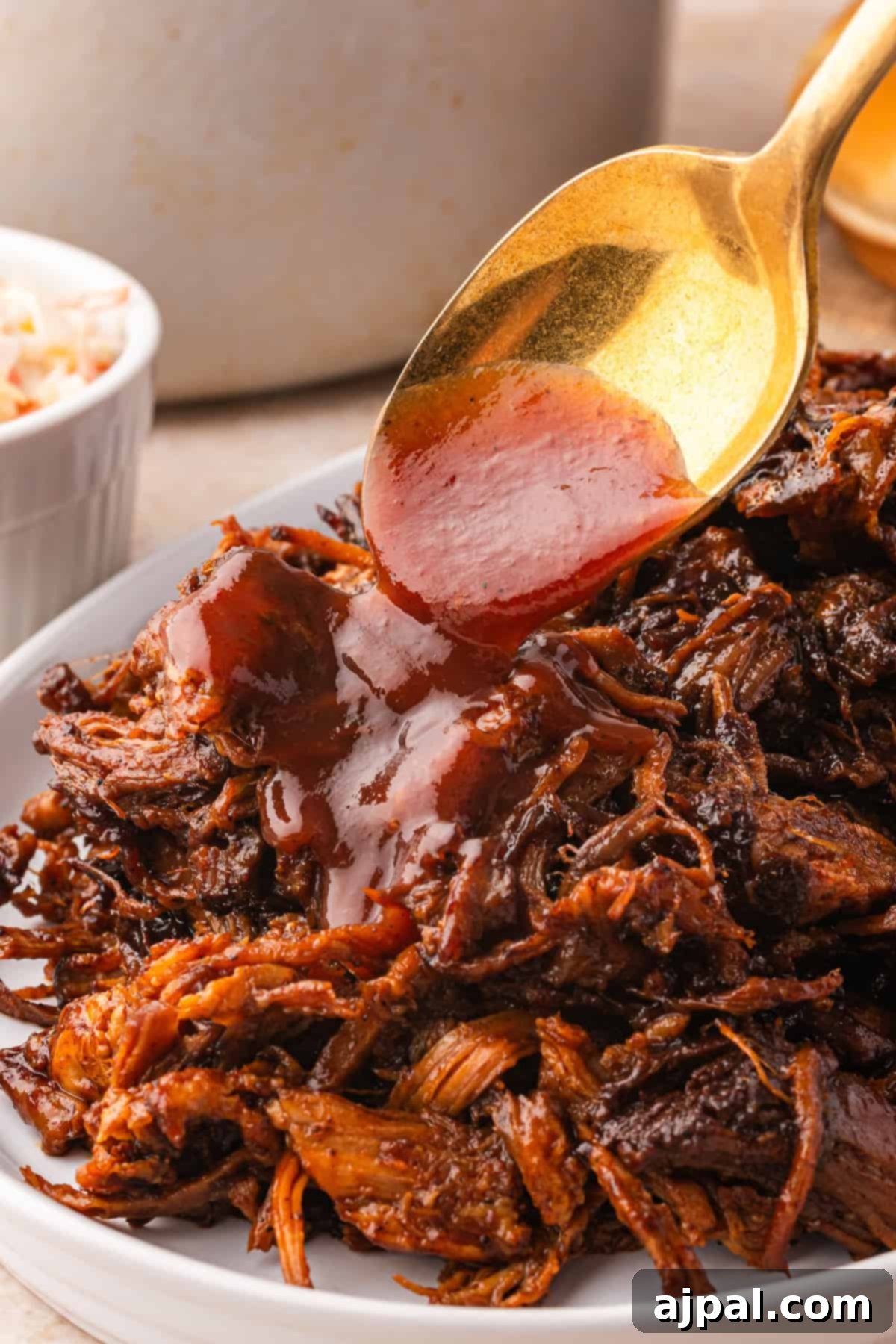 A spoon drizzles barbecue sauce over a plate of tender pulled pork, with a small bowl of coleslaw in the background, hinting at the slow-cooked magic that unfolded in the Dutch oven.