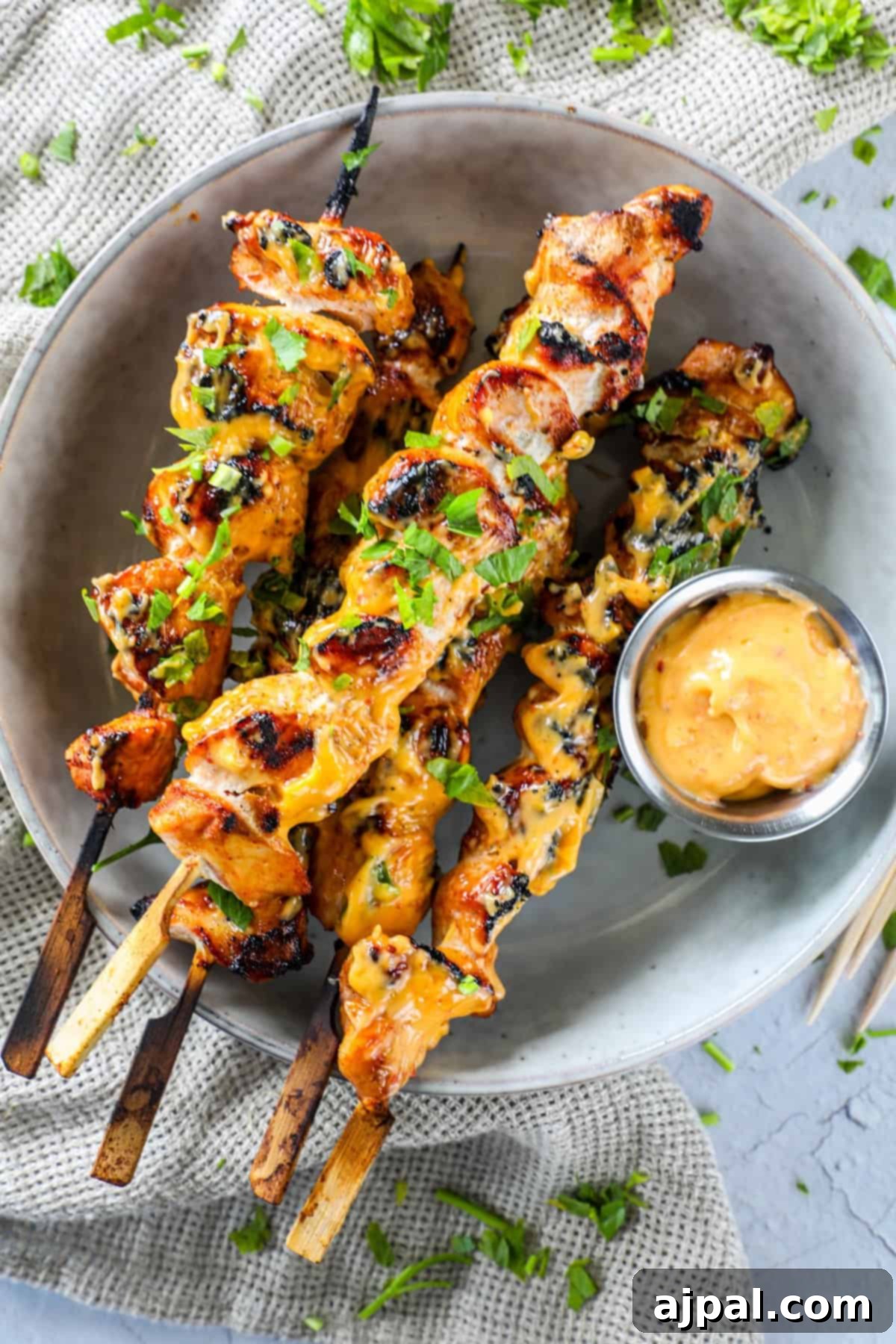 Spicy Grilled Chicken Skewers 2 Grilled chicken skewers covered in a vibrant yellow sauce, generously garnished with chopped fresh herbs like cilantro or parsley, and served alongside a small bowl of the creamy dipping sauce on a pristine white plate. The presentation highlights the saucy, herb-infused chicken.