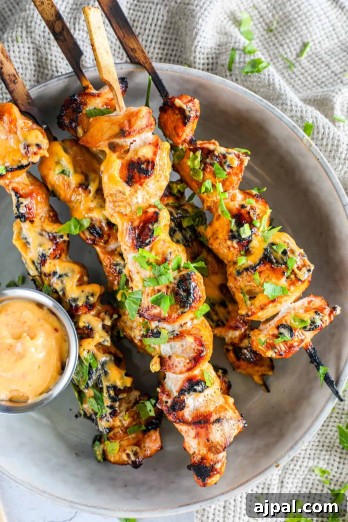 Spicy Grilled Chicken Skewers 3 Close-up of three grilled chicken skewers generously drizzled with a creamy, yellow-orange sauce, beautifully garnished with fresh chopped green herbs. A small, white ramekin holds additional dipping sauce, all presented on a rustic white plate with a subtle texture.