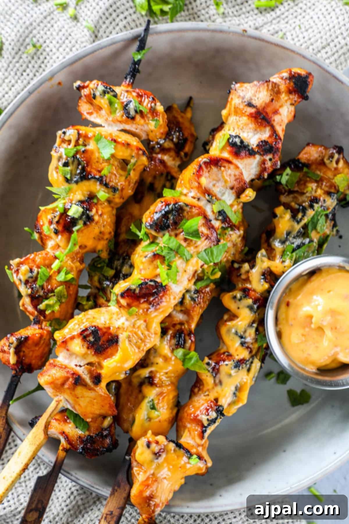 Spicy Grilled Chicken Skewers 4 Three grilled chicken skewers neatly arranged on a white plate, each generously coated with a yellow sauce and sprinkled with fresh green herbs. A small, elegant bowl of dipping sauce sits invitingly beside them, ready to enhance the flavor.