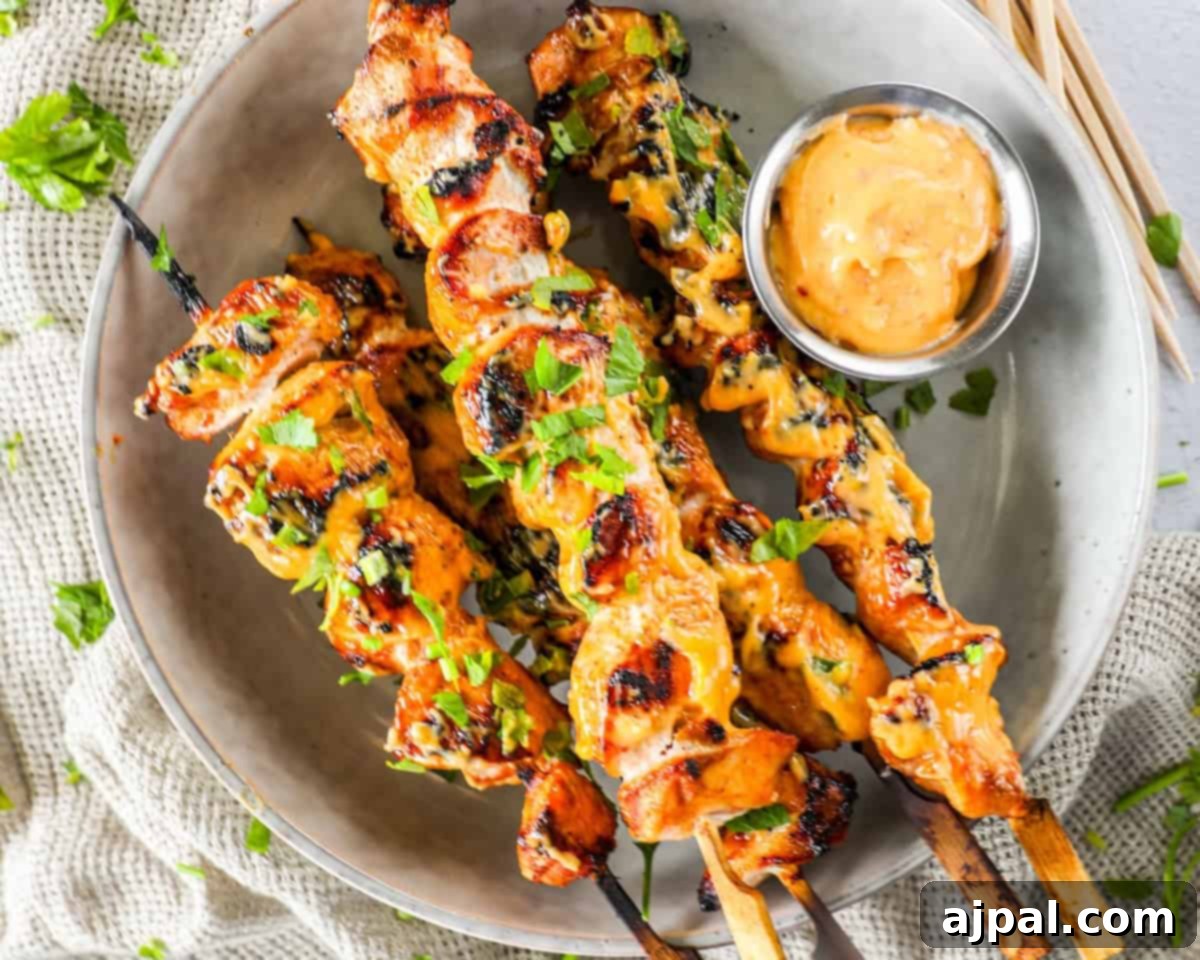 Dynamite Bang Bang Chicken Skewers 6 Plate of grilled chicken skewers garnished with parsley, accompanied by a small bowl of dipping sauce. The skewers are arranged on a beige plate placed on a textured fabric.