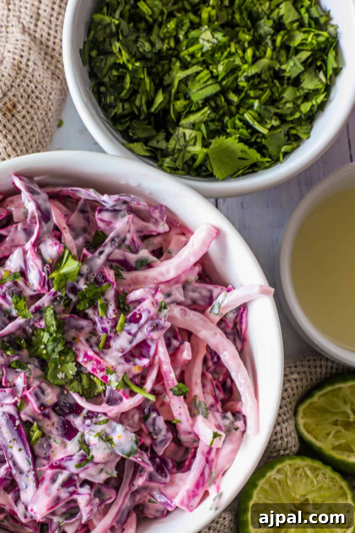 Simple Taco Slaw 2 A bowl of creamy coleslaw made with purple cabbage and garnished with herbs, alongside a bowl of chopped herbs, lime halves, and a small bowl of dressing.