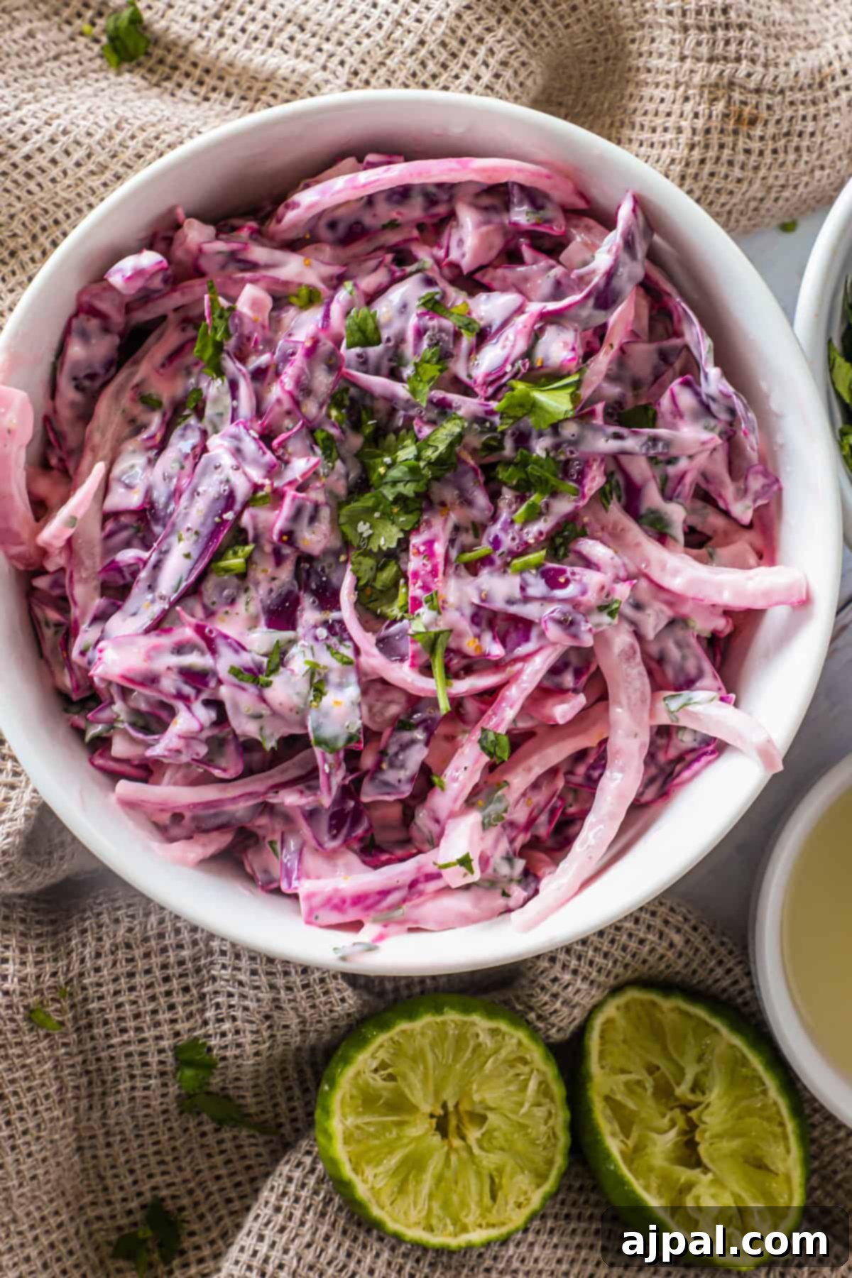 Simple Taco Slaw 9 A bowl of creamy coleslaw made with purple and white cabbage, garnished with chopped cilantro. Two halved limes are placed beside the bowl on a textured beige surface.