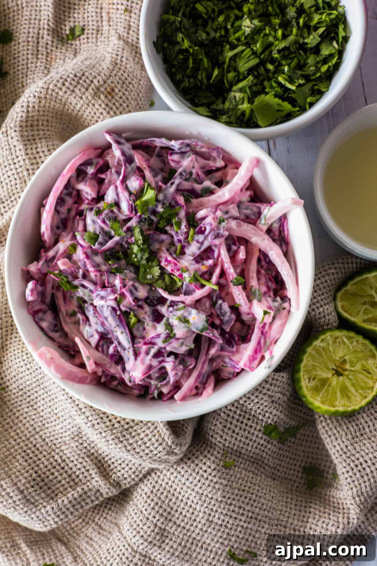 Simple Taco Slaw 10 A bowl of creamy coleslaw with purple cabbage and onions, garnished with fresh herbs, placed on a textured fabric. Bowls of chopped herbs and lime juice, as well as halved limes, are nearby.