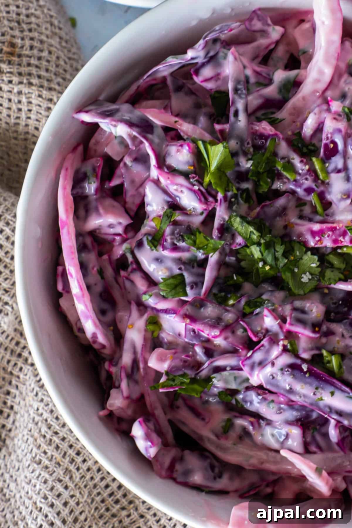 Simple Taco Slaw 4 Close-up of a bowl of creamy coleslaw with shredded purple cabbage mixed with a white dressing and garnished with chopped green herbs. The bowl is placed on a rustic fabric surface.