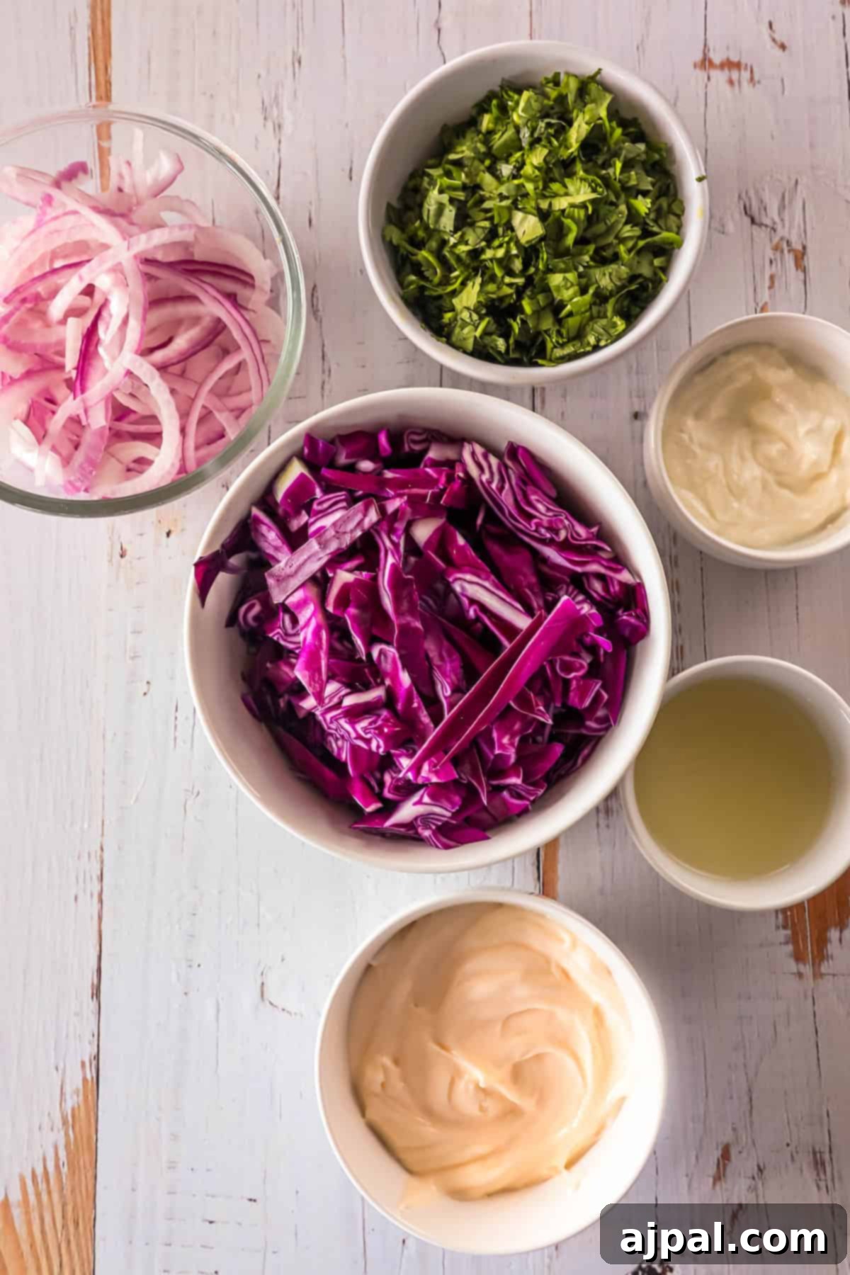Simple Taco Slaw 5 Top view of coleslaw ingredients arranged in bowls on a white wooden surface. Ingredients include red cabbage, red onion, parsley, mayonnaise, lemon juice, and a creamy dressing.