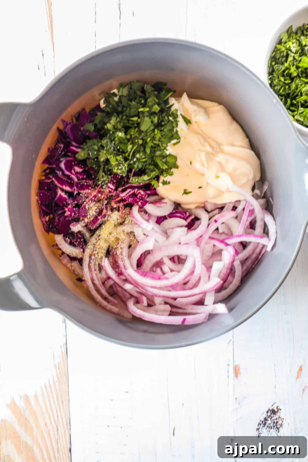 Simple Taco Slaw 6 A mixing bowl containing shredded red cabbage, sliced red onions, chopped parsley, mayonnaise, and seasonings, prepared on a light wooden surface with additional chopped parsley in a small bowl nearby.