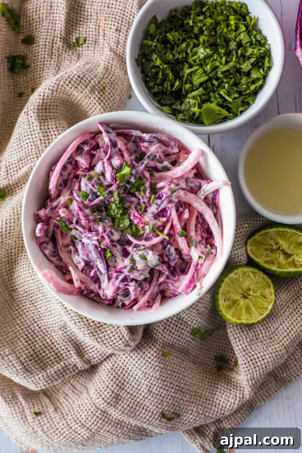 Simple Taco Slaw 7 A bowl of coleslaw with red cabbage and green herbs on a cloth, accompanied by a bowl of chopped herbs, a cut lime, and a cup of light-colored liquid.